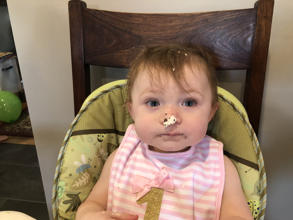 A young baby girl sitting in a high chair covered in what looks like birthday cake and frosting